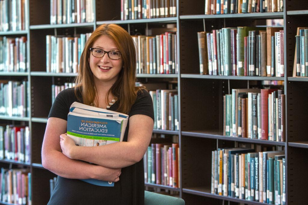 A woman with glasses stands in a library, smiling and holding a stack of books, including one titled "Foundations of American Education." She is dressed in casual attire.